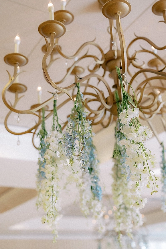 Hanging floral decor with wedding chandelier flowers, cascading white and blue delphinium, greenery, taper candles, and crystal drops indoors