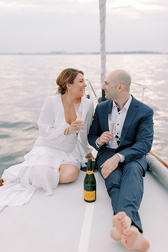 Couple portrait of bride and groom on boat laughing on a sailboat deck, holding champagne flutes with the water and horizon behind them
