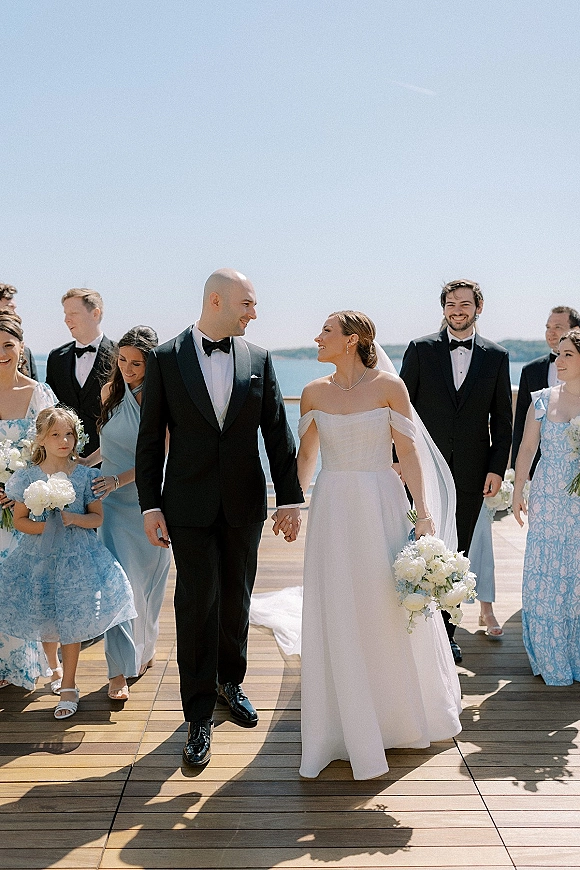 Wedding party portrait of bride and groom walking with bridesmaids in blue dresses and tuxedoed groomsmen on a waterfront deck, bouquet in hand
