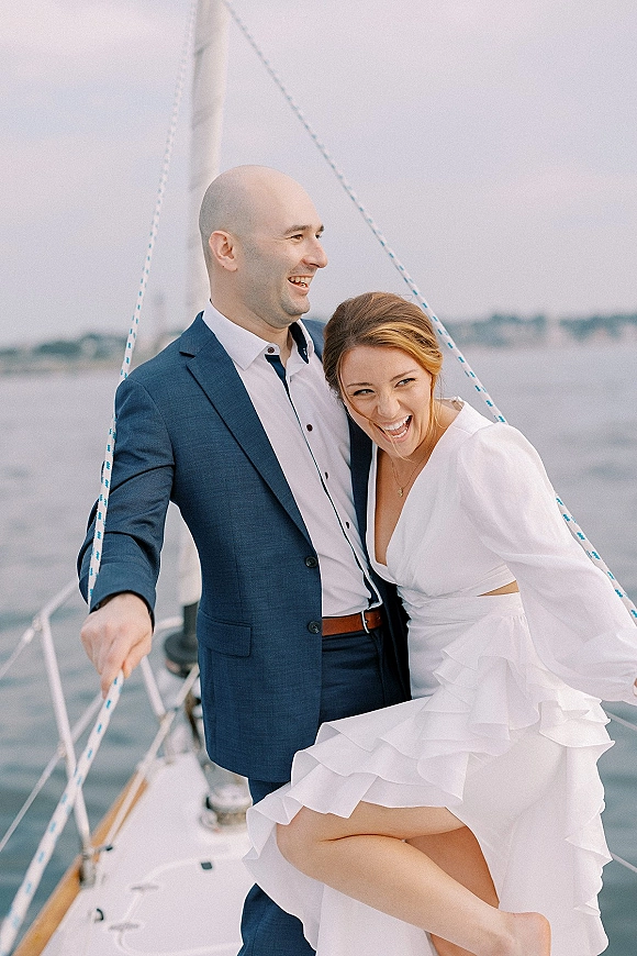 Couple portrait on a sailboat wedding portrait, laughing in a white bridal dress and blue suit on deck with ropes, mast, and water backdrop