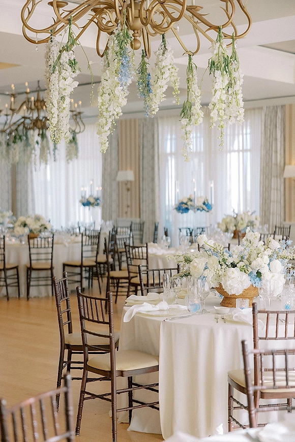 Reception tablescape with round wedding reception tables dressed in white linens, blue floral centerpieces, taper candles, and chandelier in a ballroom