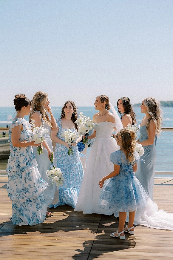 Bride with bridesmaids holding bouquets in blue dresses beside her veil and strapless gown on a waterfront deck under blue sky