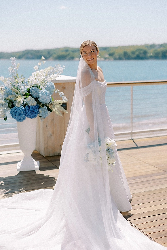Bridal portrait of a bride with veil holding a white and blue hydrangea bouquet on a wooden deck overlooking a lake and hills