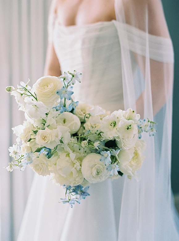 Bridal bouquet of white and blue wedding bouquet blooms tied with ribbon, held against a strapless dress and veil before soft curtains indoors