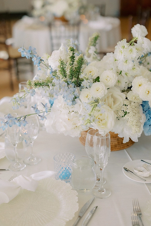 Wedding tablescape with a white and blue wedding centerpiece of white roses and delphinium on white linens, candles and glassware in reception room