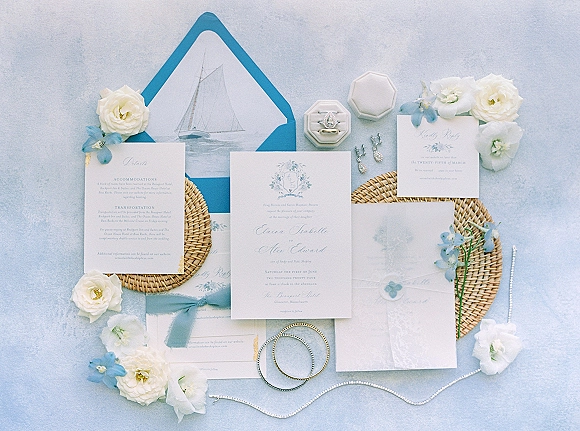 Invitation suite wedding invitation flat lay with blue envelope liner, sailboat card, wax seal, rings, pearl jewelry and flowers on light blue surface