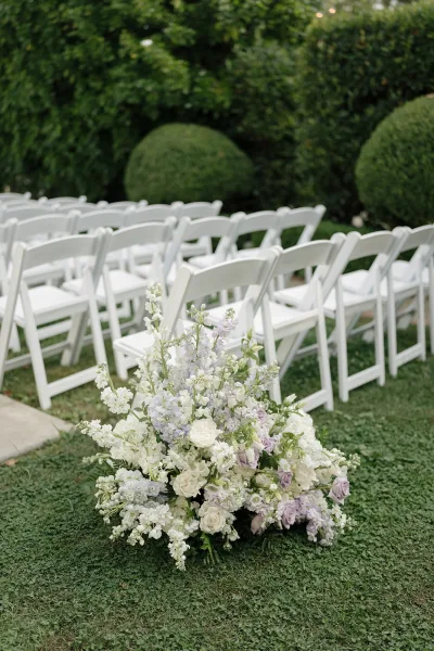 Ceremony aisle florals with lavender and white roses and greenery lining white folding chairs on a garden lawn with hedges and trees