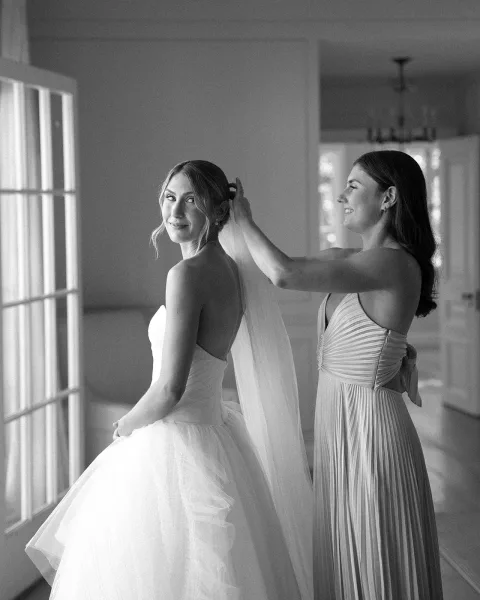 Bridal getting ready as a bridesmaid puts on veil over the bride’s updo, strapless tulle gown in a bright room with french doors