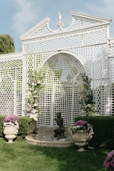 Wedding ceremony backdrop with a white lattice wedding arch and pastel floral arrangements, flanked by urn planters on a garden lawn