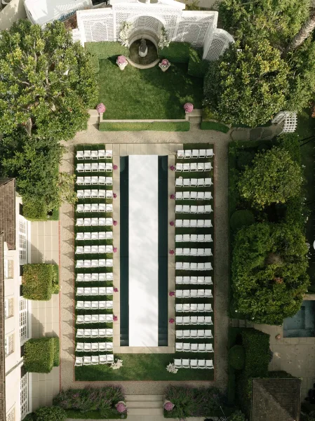 Ceremony setup with outdoor ceremony seating arranged symmetrically around a white aisle runner, leading to a fountain in a garden courtyard