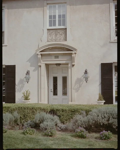 Wedding venue exterior with white double doors, black shutters and wall lanterns, framed by manicured hedges and potted plants on a stucco facade