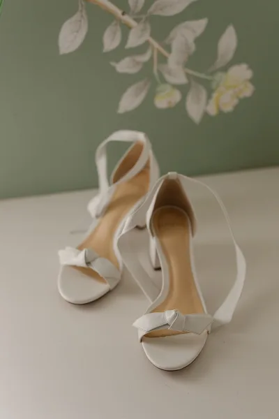 Bridal shoes, white wedding heels with strappy open-toe design on a tabletop against a green wall with floral wall art