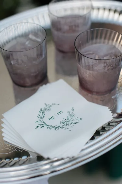 Wedding cocktail napkins with monogram cocktail napkins on a silver tray beside purple tumblers of iced drinks, set against outdoor greenery