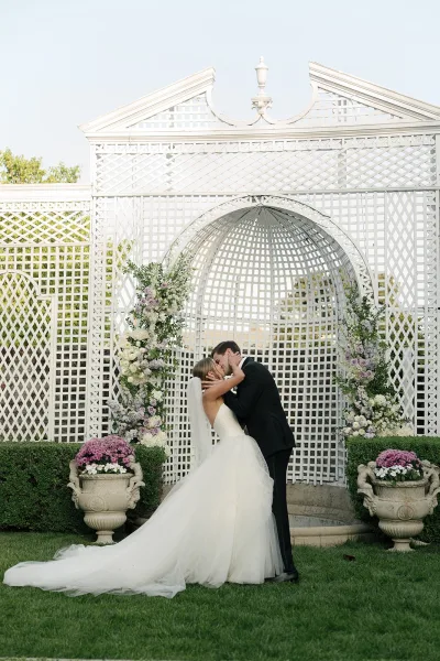 Wedding kiss portrait of bride and groom kissing under a floral arch, her veil trailing over the lawn beside white lattice trellis backdrop