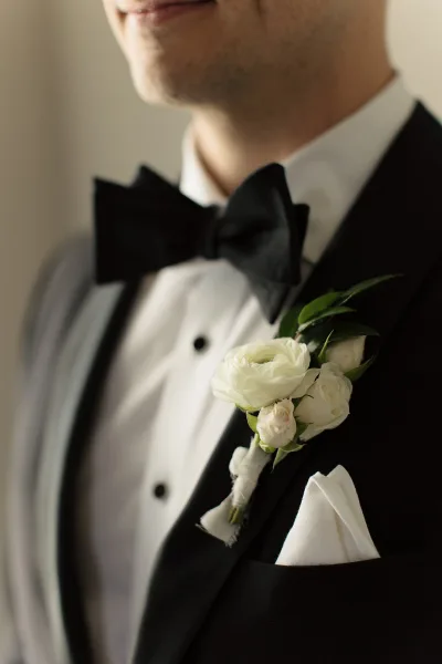 Groom boutonniere with white rose and greenery pinned to a black tuxedo lapel beside a crisp pocket square in soft indoor light