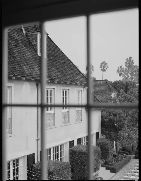 Wedding venue exterior viewed through window panes, showing stucco villa with shingle roof, balcony railings, courtyard chairs, hedges, and palms