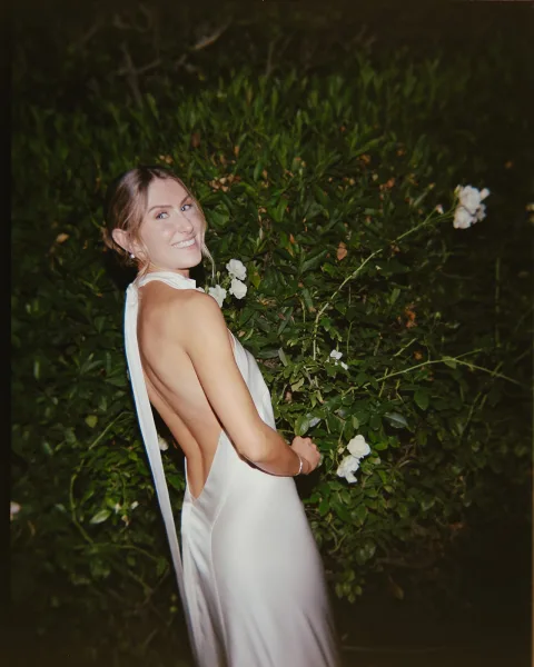 Bridal portrait of a bride in a backless wedding dress, smiling over her shoulder at night amid green foliage and white flowers