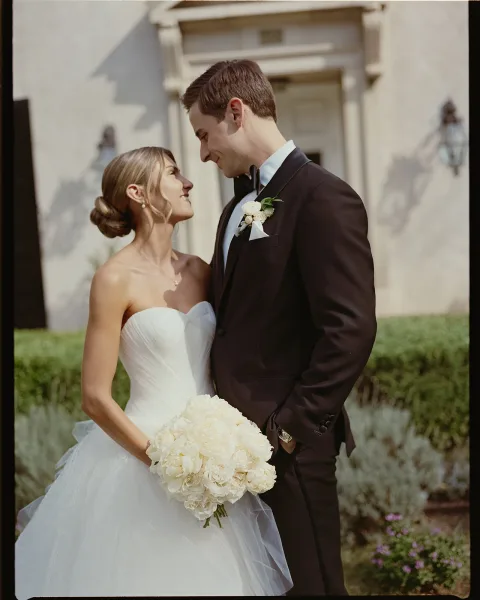 Couple portrait of bride holding a white rose bouquet and groom in black tuxedo, smiling in a garden by a doorway with hedges