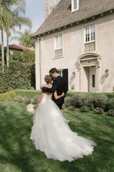 Couple portrait of bride and groom embracing, her tulle train and hydrangea bouquet flowing on a garden lawn with palms behind