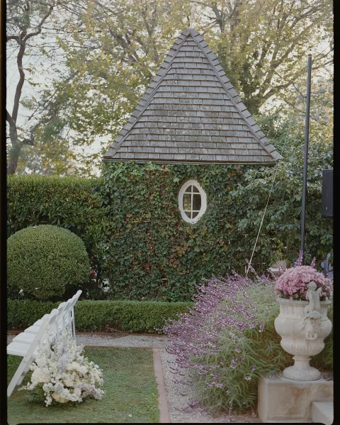 Outdoor ceremony setup with garden ceremony chairs lining a gravel aisle, featuring a stone urn of purple flowers by an ivy-covered wall