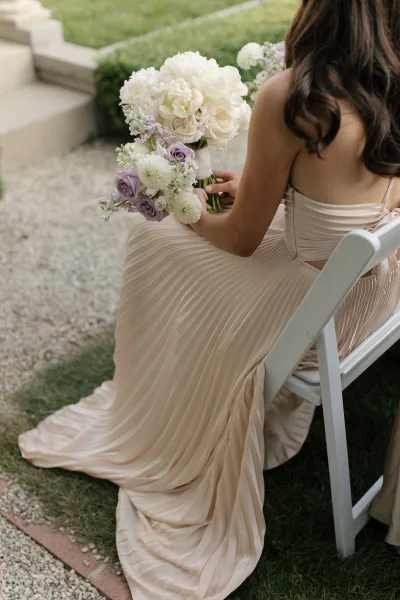 Bridesmaid holding bouquet, a pastel wedding bouquet of white peonies and lavender roses with ribbon wrap, seated by a gravel path on grass lawn