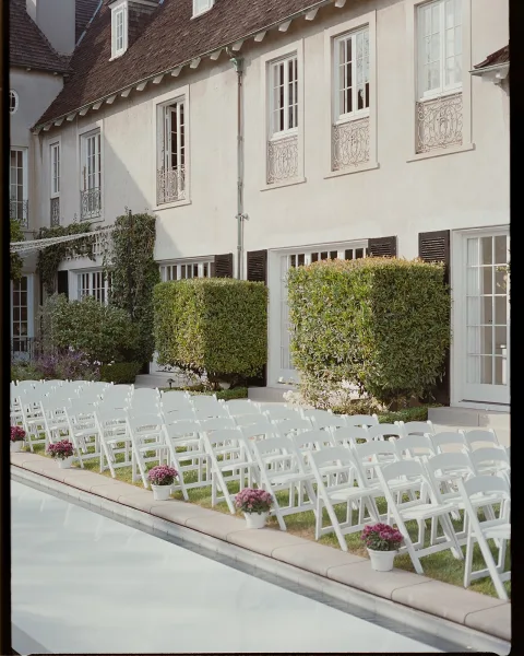 Ceremony seating with white folding chairs arranged poolside on a lawn, potted flowers marking the aisle beside a stucco villa