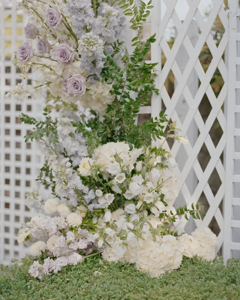 Wedding altar flowers with ceremony arch flowers in lavender roses, white blooms, and greenery cascading over white lattice panels outdoors