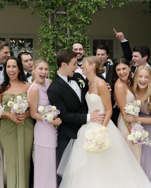 Wedding party photo of bride and groom with bridesmaids laughing, holding white and lavender rose bouquets before a vine-covered house facade.