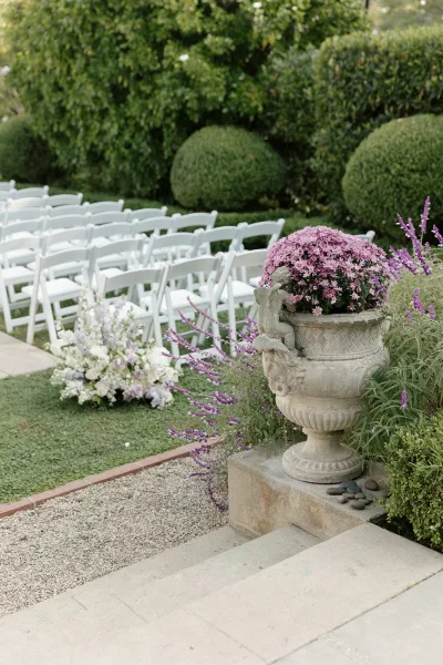 Ceremony seating with white folding chairs lining a garden lawn aisle, featuring a low purple and pink floral arrangement near stone steps