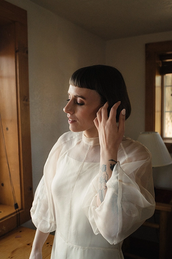 Bridal portrait of a bride getting ready in a sheer puff-sleeve, high-neck dress, touching her bob haircut by window light near a wooden wardrobe