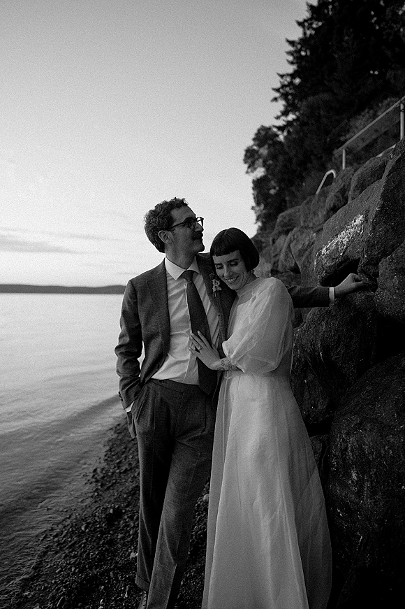 Couple portrait in a black and white wedding portrait style, bride in long-sleeve dress leaning on groom in suit with glasses on rocky shore.