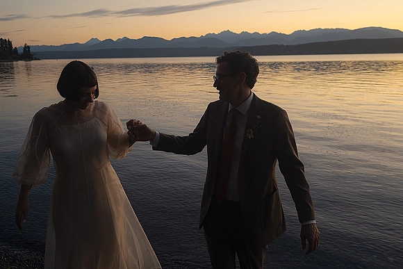 Couple portrait of newlyweds holding hands on a lakeshore at sunset, bride in sheer puff-sleeve dress and groom in suit with glasses.