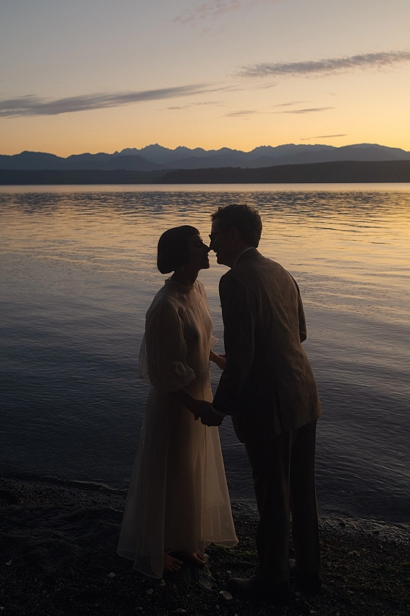 Wedding couple portrait at a mountain lake shoreline at sunset, bride in long sleeve dress and groom in light suit holding hands to kiss