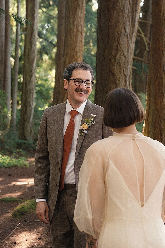 First look moment as groom in brown suit and glasses holds bride in sheer-sleeved dress on a woodland path among tall trees