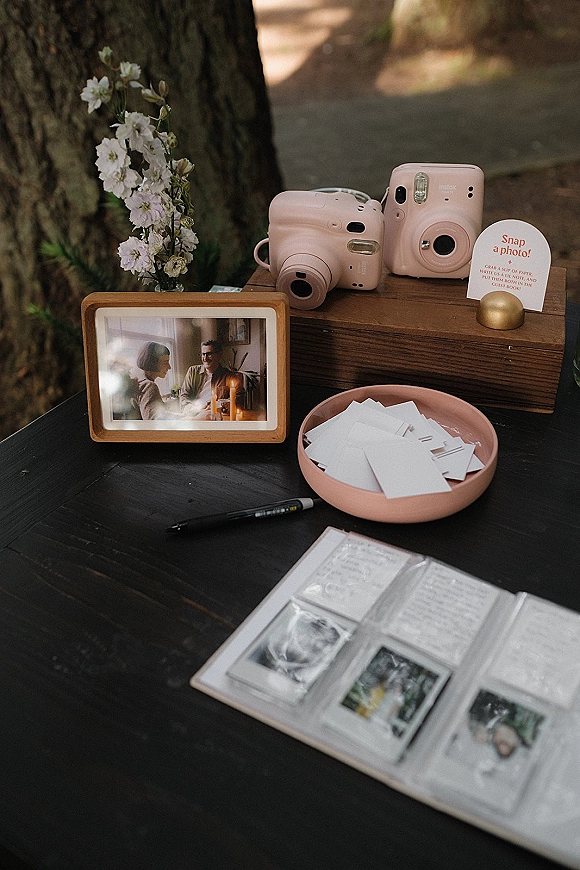 Wedding guest book with instant camera guest book setup, pen and photo slips beside a wooden box and framed photo on an outdoor table
