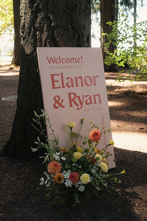 Wedding welcome sign printed on a board with a wildflower arrangement and greenery at the base, set against a sunlit forest tree trunk