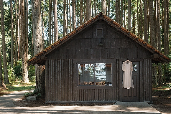 Wedding dress hanging from a dress hanger on a cabin door, high neck and short sleeves, with forest trees and window behind