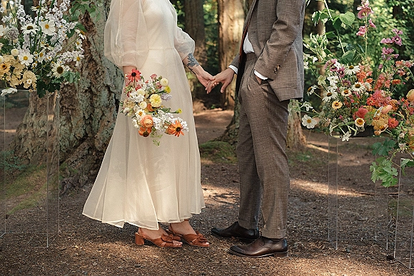 Ceremony moment with bride and groom holding hands, bouquet accents, and wildflower arrangements on acrylic stands along a forest path