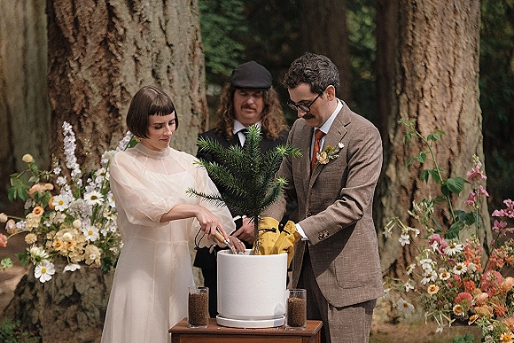 Wedding ceremony ritual as bride and groom plant a potted tree at a small table in a forest setting, with colorful florals behind them