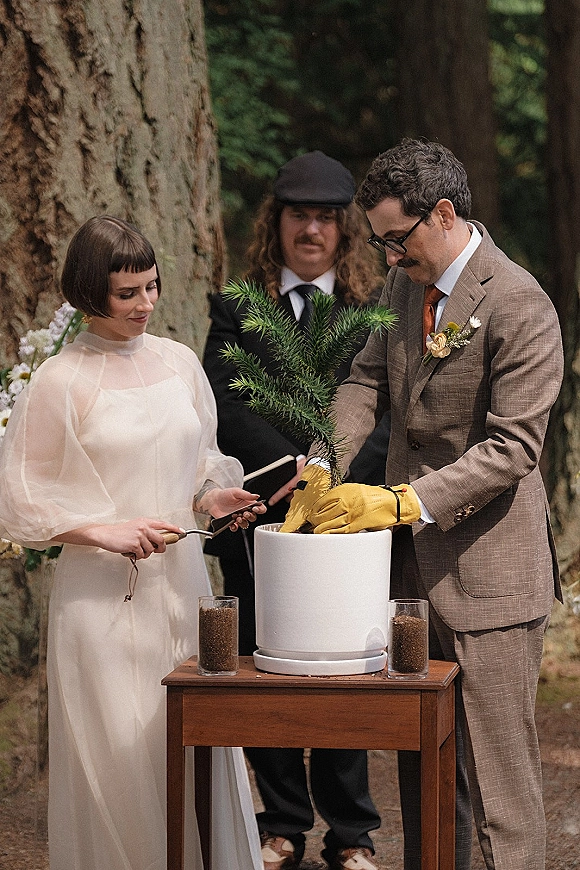 Wedding ceremony moment as bride and groom in gloves plant a tree in a pot on a wooden table, with forest trees behind them