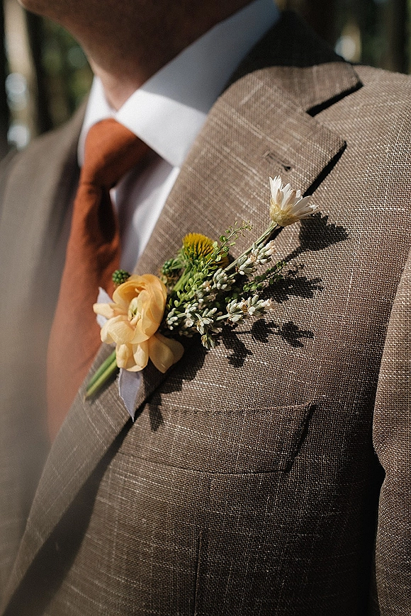 Wedding boutonniere pinned to a tan suit jacket with a burnt orange tie, crisp white shirt, and soft greenery background in sunlight