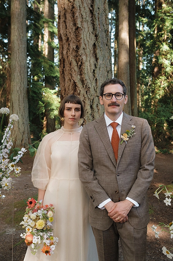 Couple portrait outdoor wedding portrait of bride in sheer-sleeve dress holding a wildflower bouquet beside groom in glasses on a forest path