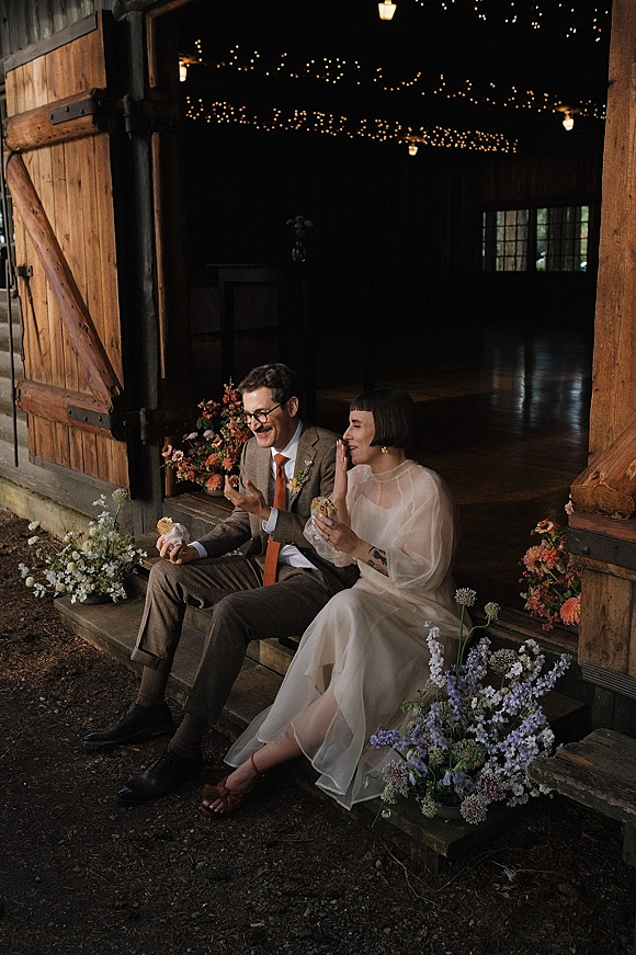 Couple portrait of newlyweds sitting on wooden steps, laughing with cookies in hand under string lights by rustic barn doors