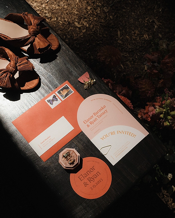 Invitation suite with terracotta wedding invitations, save the date and RSVP cards, rings and earrings on a dark wood table with florals