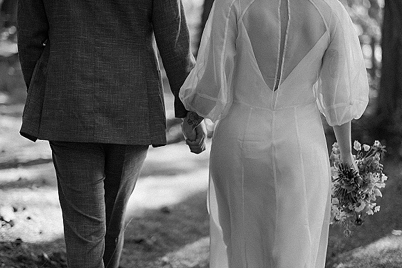 Couple portrait of bride and groom holding hands from behind, her open-back long-sleeve gown and bouquet in a sunlit garden