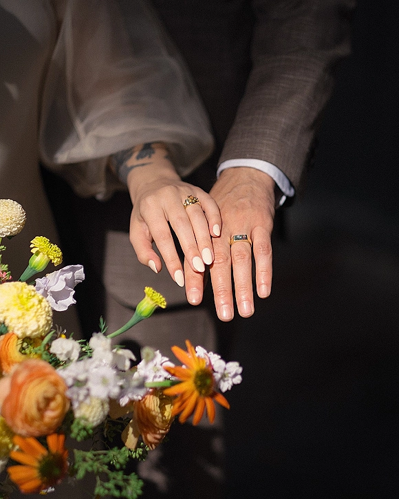 Wedding rings and engagement ring close up on a couple’s hands with nude manicure, suit jacket and veil tulle against a neutral backdrop