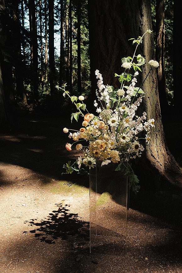 Ceremony aisle flowers in a wildflower arrangement on a clear acrylic stand, featuring daisies, blush ranunculus, and greenery in a sunlit forest