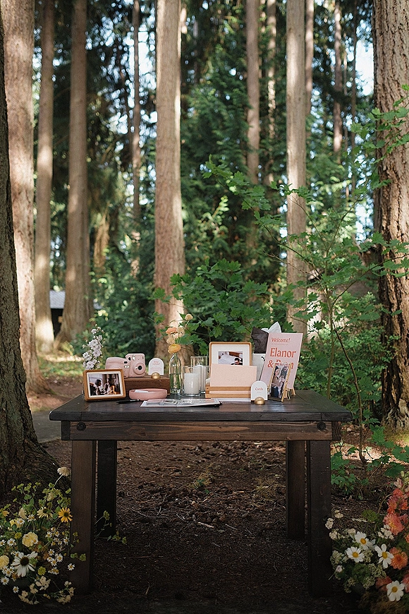 Welcome table decor with wedding welcome table sign, framed photos, instant camera guest book, candles, bud vases and flowers on a wooden table in a forest path