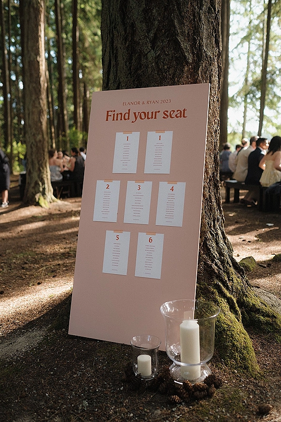 Wedding seating chart and find your seat sign taped to a board by a tree, with pillar candles in glass hurricanes and pinecones in a forest setting
