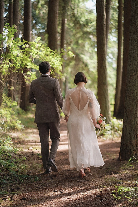 Couple portrait of bride and groom walking away hand in hand on a woodland trail, bride holding bouquet in a sunlit forest